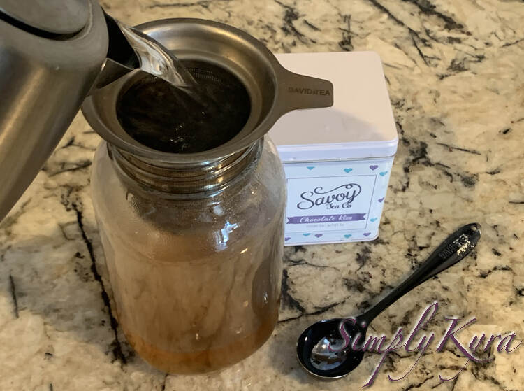 Image shows the metal kettle pouring water into the metal infuser inside the canning jar's opening. Behind and beside it sits a closed tin of tea alongside a tea measuring spoon. 