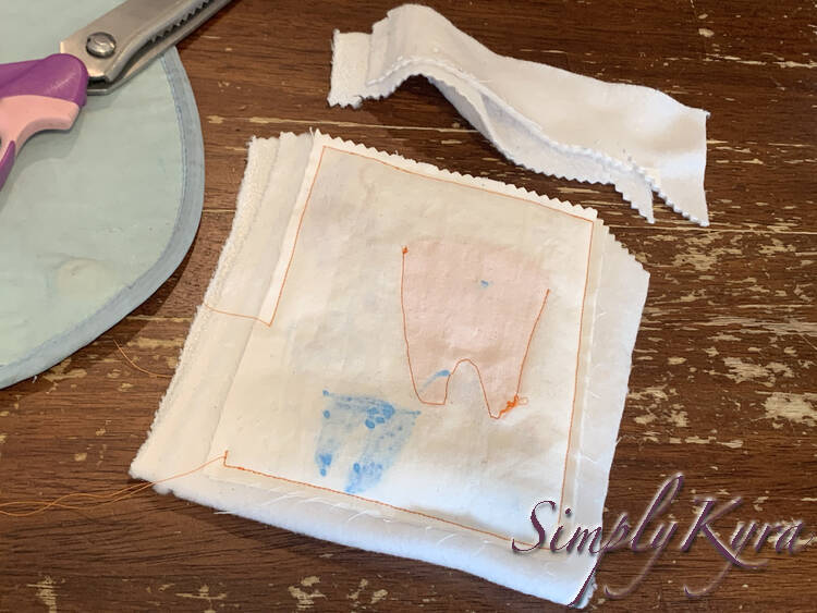 Image shows the backside of Zoey's tooth fairy where you can see the orange thread around the entire thing and around the tooth. You can faintly see the orange toothed fabric and the blue fabric marker on the other side of the thin fabric. The ironing mat and pinking shears lay to the left and the top is trimmed off and laid above. 