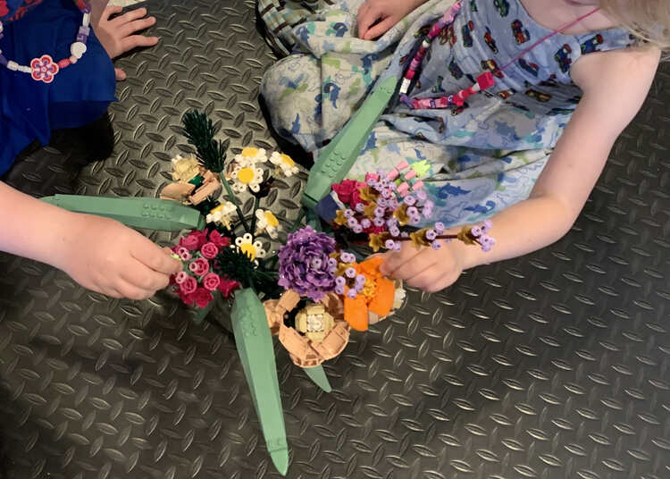 Image shows an overhead photo of the bouquet spread out in a large vase over a black floor mat. Two girls are seen to the left and right both touching a particular favorite flower. 