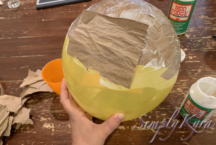 Image shows me holding a yellow balloon above the table with the top coated in brown paper and white streaks. The front has a dry brown piece of paper mostly sticking to it. 