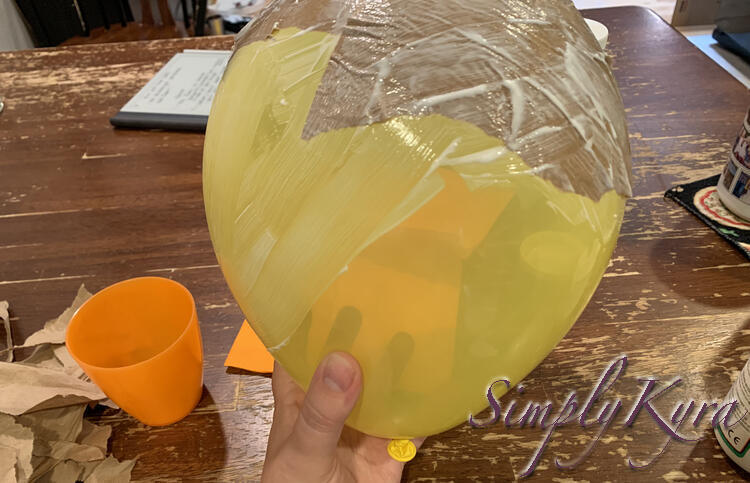 Image shows me holding a yellow balloon above the table with the top coated in brown paper and white streaks. The front has a lot more Mod Podge streaks over the yellow balloon.