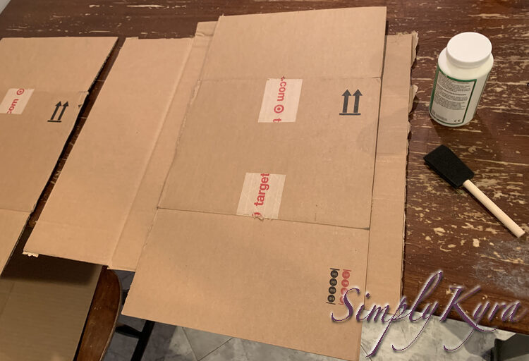 Image shows one larger square of cardboard underneath with a smaller rectangular Target box covering the right side. The other piece is to the left while a bottle of Mod Podge and a foam brush lay to the right. 