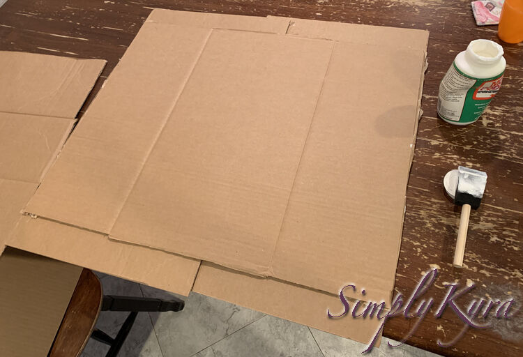 Image shows the same vague rectangle of cardboard but this time it's three layers tall with the smaller Target boxes peeking out from the front and back. The almost empty jar of Mod Podge and the wet brush sit to the right side of the photo. 