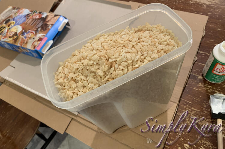 Image shows the same cardboard but this time the  image is taken from higher up and a plastic container filled with Rice Krispy cereal sits on top. And empty cereal box sits to the side on top of the emptied and flattened one from before. 