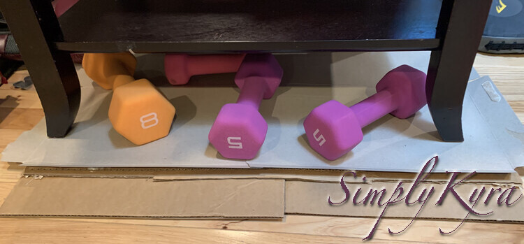 Image shows the cardboard laid out on a wood flour with a wooden piece of furniture with four legs on top of it and some bright orange, purple, and pink weights on too. 