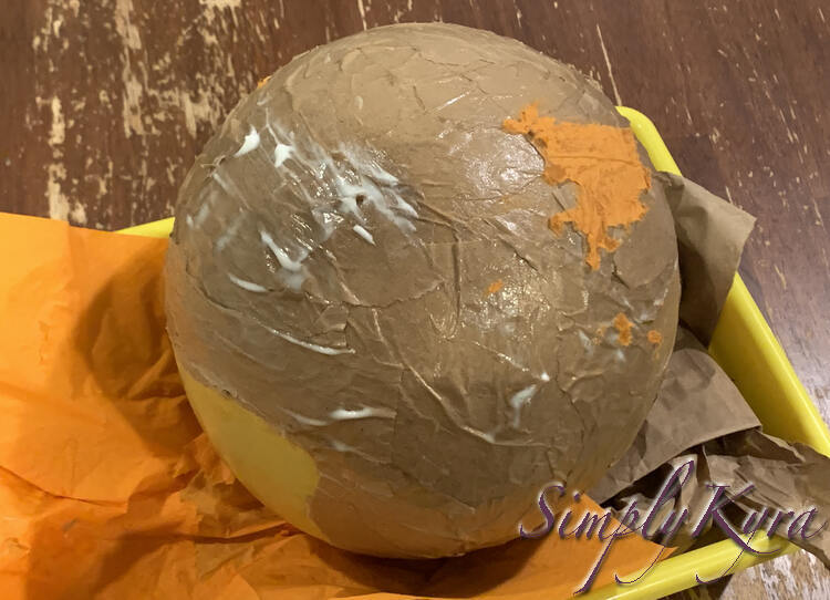 Image shows the balloon with the brown and orange encrusted side up and the yellow knotted side down. The top has white spots showing the Mod Podge hasn't dried yet there. 