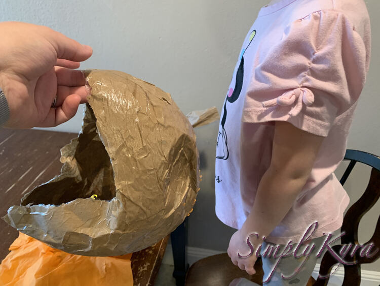 Image shows me holding the slightly crumbled helmet. The background of the image shows the discarded orange napkin on the table and Zoey standing on a chair waiting to try the helmet on.