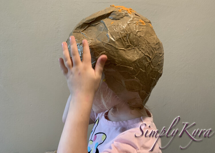 Image is a side view of Zoey wearing the orange topped brown helmet. Her hand up going up to see how it feels.