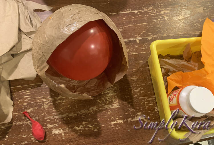 Image shows the helmet with a half sized balloon inside. Off to the side is the paper, the Mod Podge, the ripped napkin, and another red balloon waiting to replace the second one inside the helmet. 