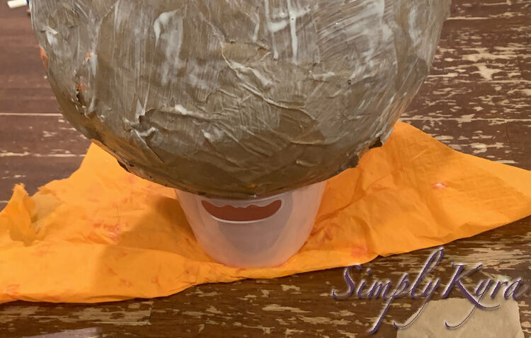 Image is a side view of the pink basket holding the wet white streaked balloon above the table and orange napkin. 