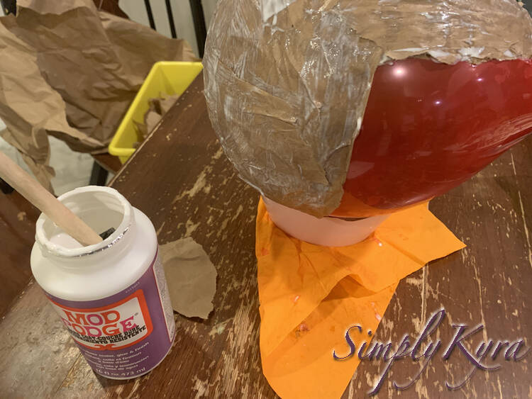 Image is a side view of the previous photo. This time there's an opened bottle of Mod Podge with a foam brush inside beside it and you can see the yellow bin of paper in the chair behind.