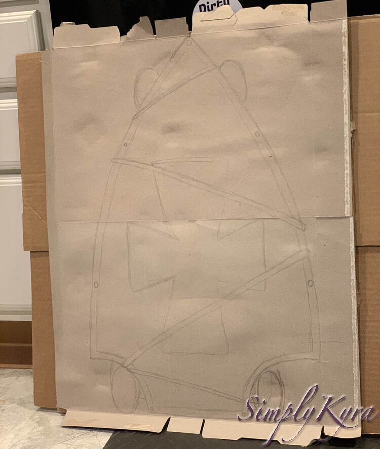 Image shows the cardboard leaned up against my cupboards on the kitchen floor. On it you can see the skateboard sketched out showing the shield shape, it's details, the four wheels, and the lines were the necklace was used to attach them together. 