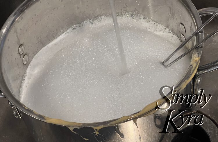 Image shows a metal pot with brown honeycomb candy stuck to the edge, a wire whisk handle sticking out, and soapy water being filled inside. 