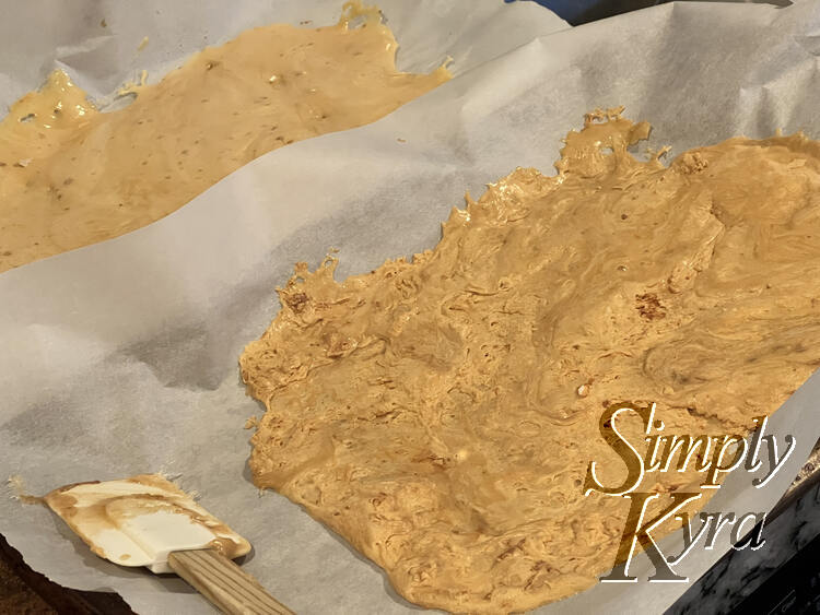 Image shows two trays of honeycomb candy side by side on parchment lined cookie sheets. Beside them rests a candy coated silicone spatula. 