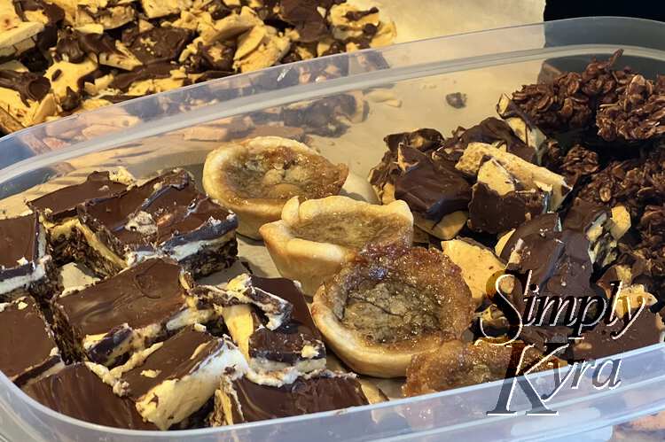 Image shows a large clear plastic container with stacks of Nanaimo bars, parchment separated butter tarts, chocolate coated honeycomb candy, and chocolate haystacks. In the background rest the remaining honeycomb candy. 