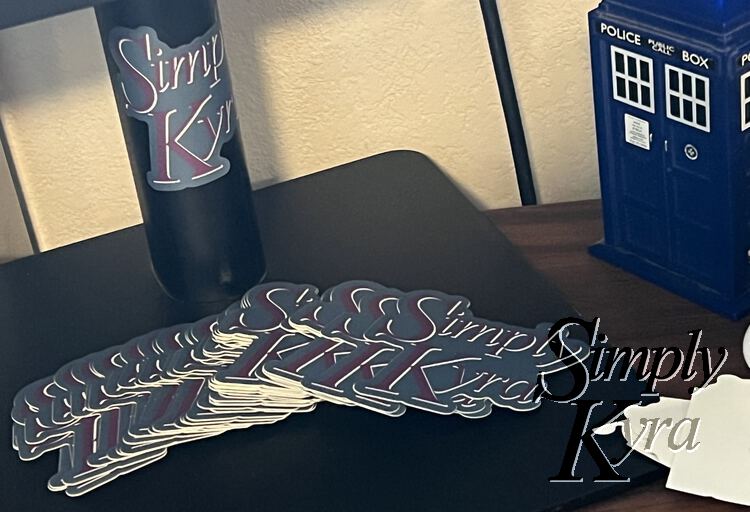 Image shows the pile of custom SimplyKyra stickers spread out on the base of my computer monitor's stand. In the center is one stuck to the center stand. A plastic USB TARDIS sits off to the side.  