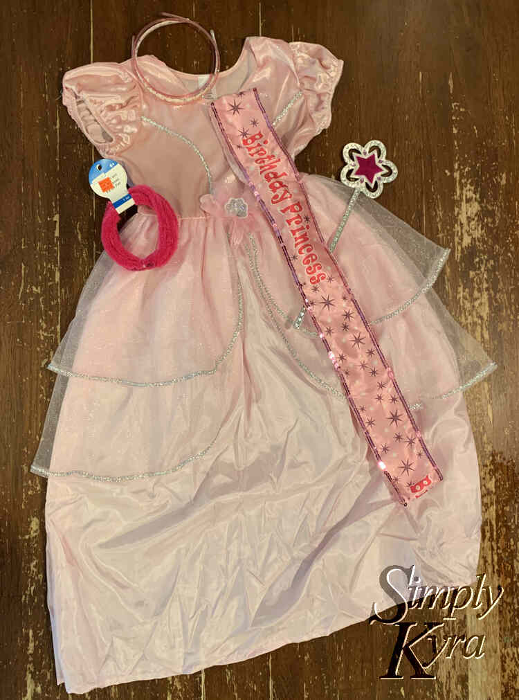 Image shows the dress laid out on the kitchen table with the headbands, pink chenille strand, wand, and birthday sash laid out over and around it. 