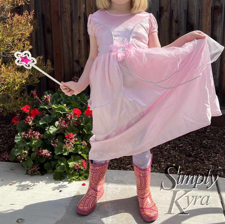 Image shows Ada standing in pink rubber boots wearing the pink glittery dress. One hand is holding the skirt high showing her pink hearted pants while the other holds the wand.