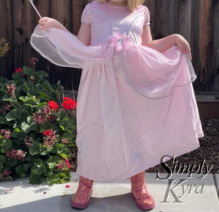 Image shows Ada standing in pink rubber boots wearing the pink glittery dress. One hand is holding the skirt up while the other holds the wand and the tulle overskirt. 