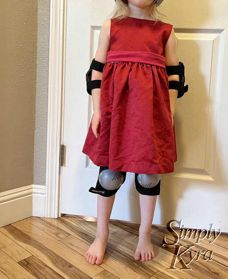 Zoey standing barefoot on the floor in her red dress, pink sash, elbow pads, and knee pads. 