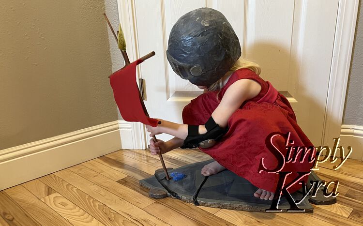 Zoey is now crouched in her dress, wearing her helmet, holding onto the mast of the sail turning the skateboard into a boat. 