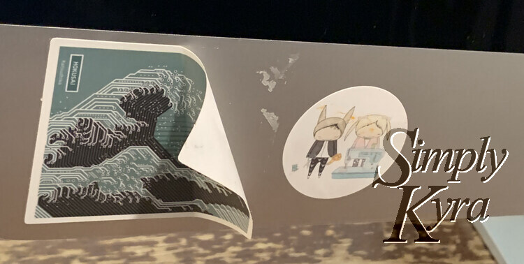 Image shows the bottom of my computer monitor. On the left is a partially peeled sticker showing a circuit board version of The Great Wave off Kanagawa. To the right is a white circular sticker with a sewing machine and two people on it from Whimsy Baby Customs.
