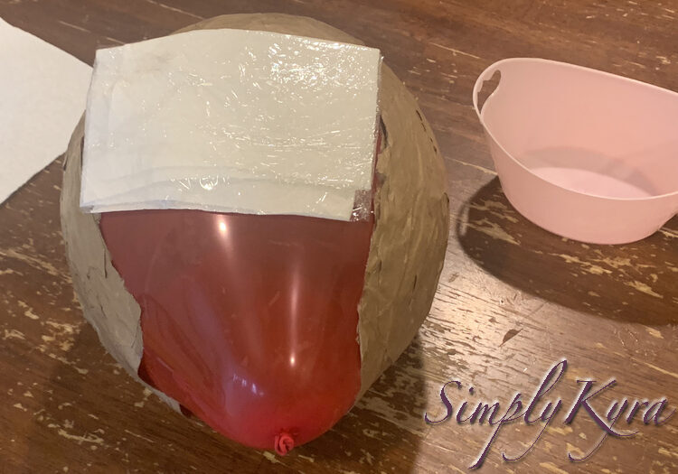 Image shows the balloon on the side so the front of the helmet is facing up. There's a rectangle of shiny white laid over the top of the opening similar to if the balloon had a headache and it was a wet washcloth. More paper towel and the pink basket sit in the background. 