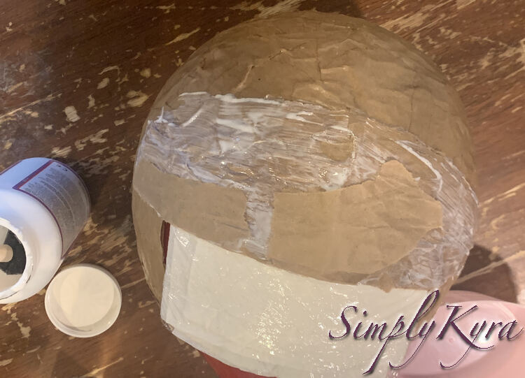 Image shows almost the same angle as before but now the vertical rolled paper is hidden away under other paper mache. The last two larger pieces that were added are apparent as they haven't been painted over yet. An open jar of Mod Podge sits to the left.
