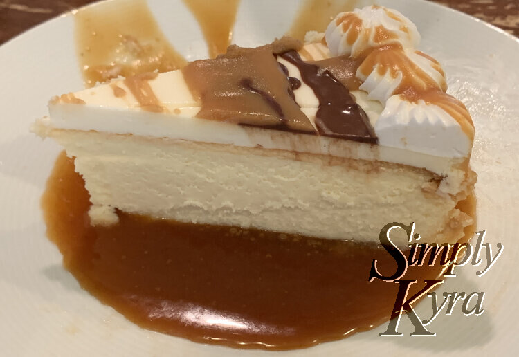 Image shows the hardened chocolate and caramel topped cheesecake slice sitting in a pool of hot caramel on a white saucer. 