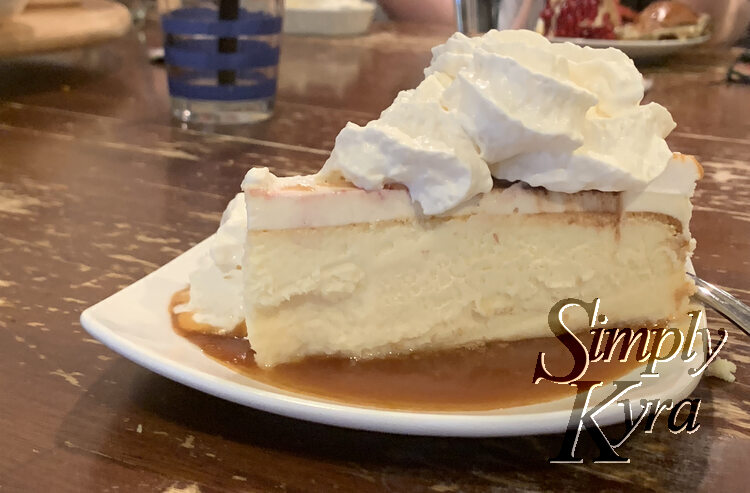 Photo is taken from the side looking at an upright slice of cheesecake sitting in melted caramel and topped with whip cream mostly hiding the leftover cold chocolate and caramel on top of the slice. 