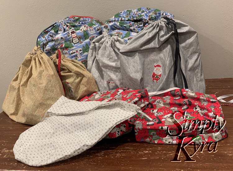 Image shows a stack of eight gifts wrapped in the fabric bags laid out on the kitchen table. An empty extra white glittered one lays in the front overlapping the cute dogs. You can see Santa or penguin labels stuck to the back ones. 