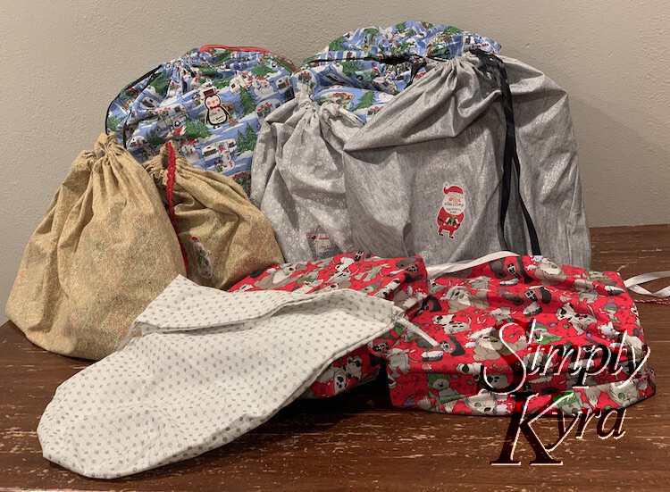 Image shows a stack of eight gifts wrapped in the fabric bags laid out on the kitchen table. An empty extra white glittered one lays in the front overlapping the cute dogs. You can see Santa or penguin labels stuck to the back ones.