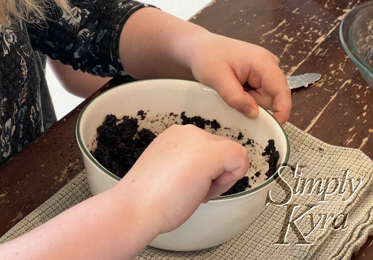 Image shows a tall white bowl on a folded tea towel. It's filled with a dirt-like consistency and someone, you can only see the arms and part of the body, is mixing it together. 