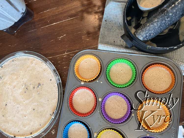 Image shows a glass pie plate filled with bubbly liquid to the left. To the right you can see six and a bit colorful cupcake liners half filled with more cheesecake mixture. To the top you can see the blender, on the left, and the blender accessories, measuring cup, and tea towel on the right. 