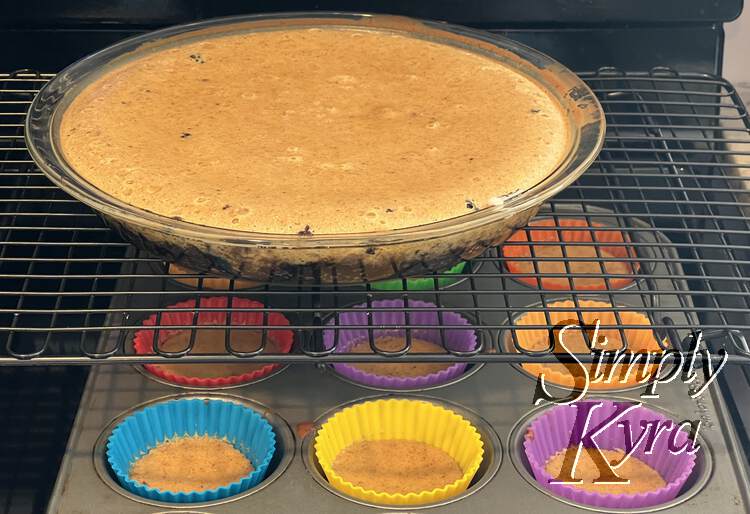 Image shows a slightly underbaked cheesecake resting on a black grilled cooling rack. Below it you can see multi-colored silicone wrapped cupcakes on the stovetop. 