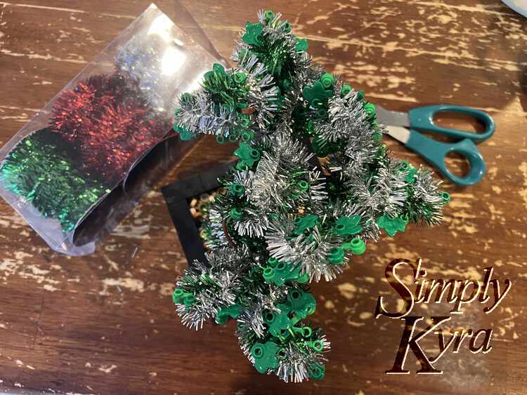 Image is taken from above showing the bonsai tree festooned with silver and green tinsel. Below and to either side is the three colored tinsel put away and a pair of scissors.