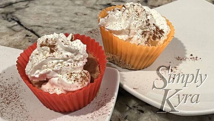 Image shows two cupcakes on two white saucers. One is in a red liner and the other orange. Both are topped with a swirl of white whip cream and a sprinkling of cinnamon.