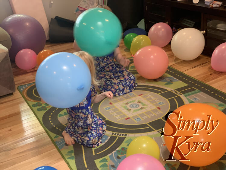 Both girls are crouched on the floor wearing their Bluey dresses and punching balloons. They're surrounded by other balloons. 