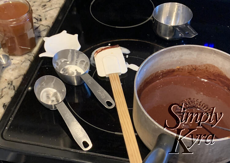 Image shows a pot of brown liquid place between the burners with dirty measuring cups and spatulas to the left of it. 