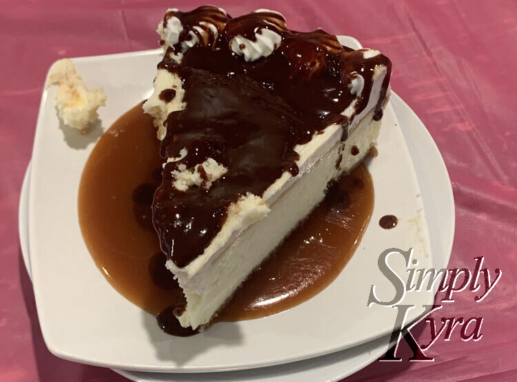 Image shows a stack of white saucers with a large slice of cheesecake sitting in a light brown pool of caramel with chocolate coating the top. Under the saucer you can see the pink tablecloth covering the table. 