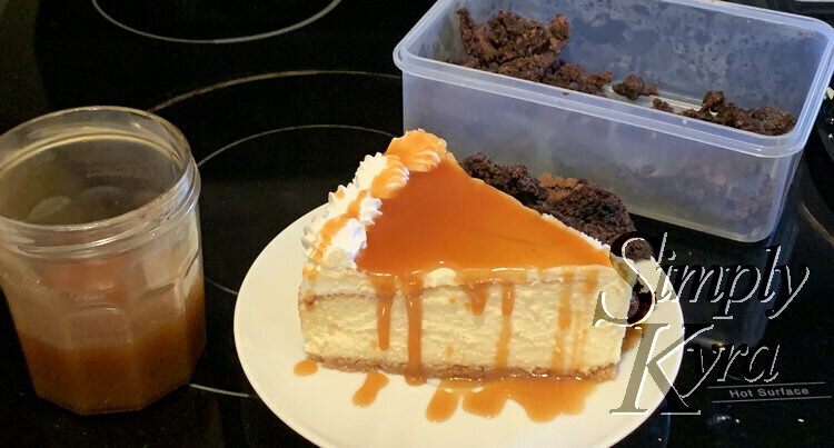 Image shows a circular saucer with a large piece of cheesecake coated in caramel with drizzles dripping down. Behind the slice you can see pieces of brownie on the saucer. Beside the saucer is a half filled jar of caramel sauce and behind it is a plastic container of brownie bits. 