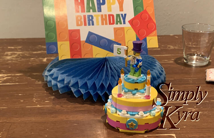 Image shows the LEGO cake with a clown holding a flag saying 5. Behind it is the Happy Birthday table centerpiece sign, an empty glass of water, and a nametag.