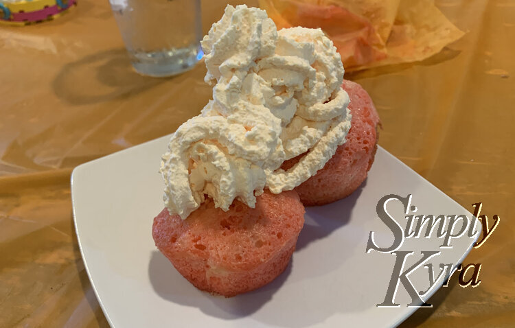 Image shows a white saucer with two pink cupcakes and tons of orange whip cream on top. The saucer sits on the orange tablecloth covered table.