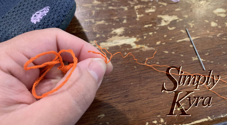 Image shows my pinching near the end of some orange thread. The end splays out and the thread on the other side is bunched up. The table below shows a freed thread along with a blunt embroidery needle.