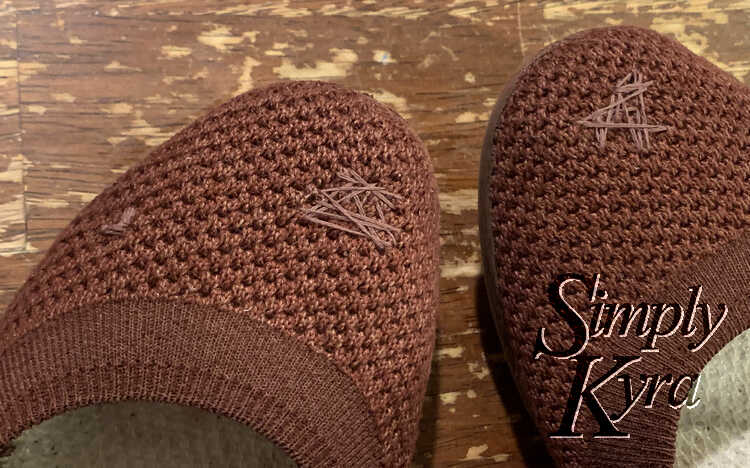 Image shows the front top of both shoes side by side over the brown table. They both have a decorative triangular or heart-ish criss-crossing design over their main hole. The left shoes also has a small left covering a little patch on the top side. 