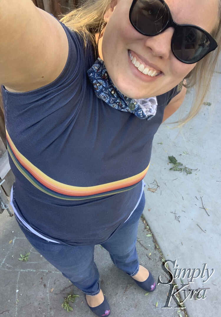 Image is taken from a high selfie angle of me smiling and looking at the camera. I'm wearing sunglasses, a blue Doctor Who face mask around my neck, a blue Doctor Who top, jeans, and my blue flats. 