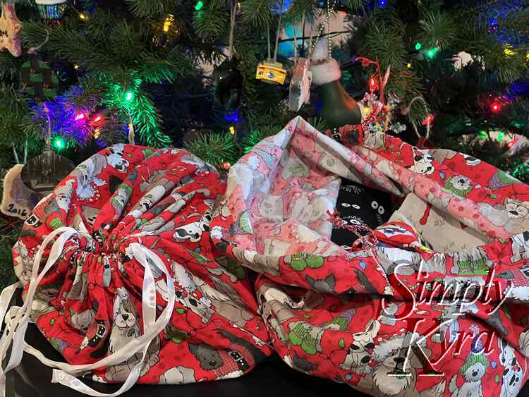 Image shows the two dog bags in front of our lit up Christmas tree. The bag on the left is cinched closed with white ribbon while the bag on the right is open showing red and eyeball covered fabric and the sparkly ribboned drawstring. 