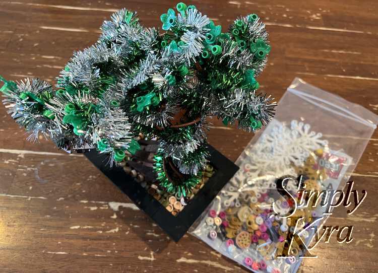 Image is taken from above looking down at the top of the bonsai draped with silver and green tinsel. To the right lays a slightly blurred clear zip locked baggie of LEGO® bricks. 