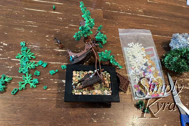 Image shows the black bonsai dish at the center of the image with the trunk broken and laid out behind the base. To the left are all the extra green pieces that fell off while, to the right, lay all the new bricks, the tinsel, and some scissors.