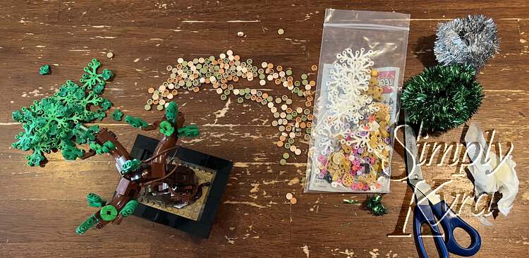 Image shows the brown trunked bonsai with a few green bricks at the end of each branch. Behind it, from left to right, lay the green bricks, the dirt, the baggie of new pieces, the scissors, rolled up tinsel, and a wet paper towel used to wipe the dust off the black base. 